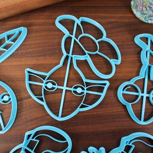 May include: Five blue plastic cookie cutters shaped like a pear, a flower, a face with eyes, and two cherries. The cutters are on a wooden surface.