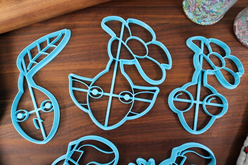 May include: Five blue plastic cookie cutters shaped like a pear, a flower, a face with eyes, and two cherries. The cutters are on a wooden surface.