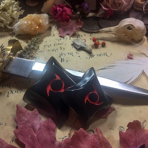 K&ouml;nnte beinhalten: Zwei schwarze, sternf&ouml;rmige Ohrstecker mit rotem Spiraldesign werden auf einem Vintage-Papierhintergrund pr&auml;sentiert. Ein silberner Dolch und getrocknete Bl&auml;tter und Blumen umgeben die Ohrstecker. Die Ohrstecker sind wahrscheinlich f&uuml;r gedehnte Ohrpiercings.