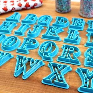 May include: A set of 26 blue plastic alphabet cookie cutters on a wooden surface. The cutters are in a decorative font style.