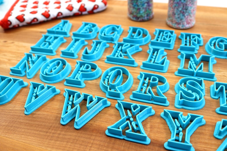 May include: A set of 26 blue plastic alphabet cookie cutters on a wooden surface. The cutters are in a decorative font style.