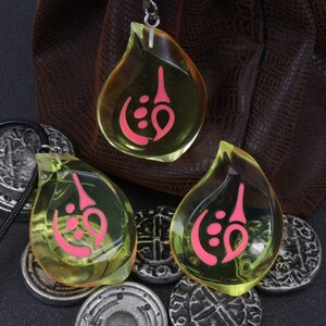 May include: Three teardrop-shaped pendants with a pink symbol inside. The pendants are made of clear yellow resin and are hanging from black cords. The pendants are sitting on a brown leather pouch and silver coins.