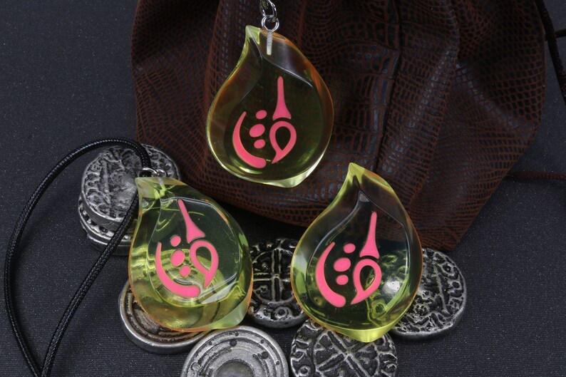May include: Three teardrop-shaped pendants with a pink symbol inside. The pendants are made of clear yellow resin and are hanging from black cords. The pendants are sitting on a brown leather pouch and silver coins.