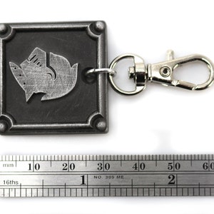 May include: A silver metal keychain with a square tag featuring a stylized wolf design. The tag has a textured surface and a silver metal clasp.