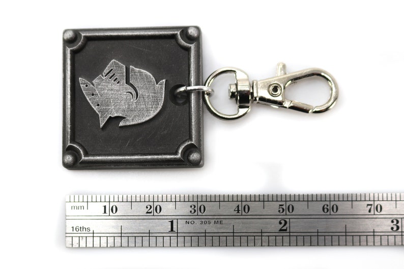 May include: A silver metal keychain with a square tag featuring a stylized wolf design. The tag has a textured surface and a silver metal clasp.