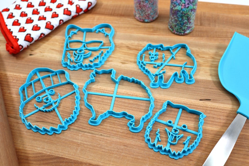 May include: Five blue plastic cookie cutters shaped like dogs. The cutters have different expressions, including sunglasses, a hat, and a tongue sticking out.