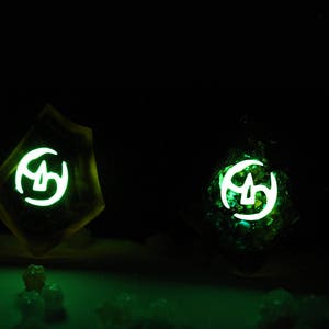 May include: Two glowing green resin dice with white symbols. The dice are on a dark surface.