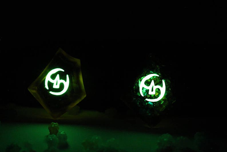 May include: Two glowing green resin dice with white symbols. The dice are on a dark surface.