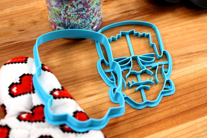 May include: Blue plastic cookie cutter shaped like Frankenstein's face. The cutter has detailed features including eyes, nose, mouth, and neck bolts.