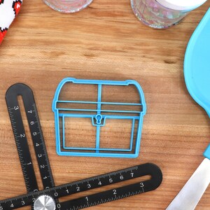 May include: A blue plastic cookie cutter in the shape of a treasure chest sits on a wooden surface. A black angle ruler and a blue spatula are also present. A white cloth with red pixelated hearts is in the background.