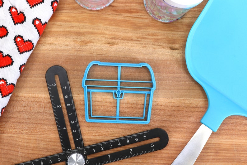 May include: A blue plastic cookie cutter in the shape of a treasure chest sits on a wooden surface. A black angle ruler and a blue spatula are also present. A white cloth with red pixelated hearts is in the background.