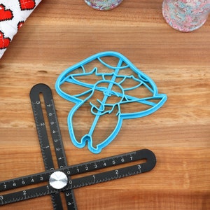 May include: A blue plastic cookie cutter shaped like a mushroom with a small mushroom on top. The cookie cutter is on a wooden surface with a ruler and other baking supplies.