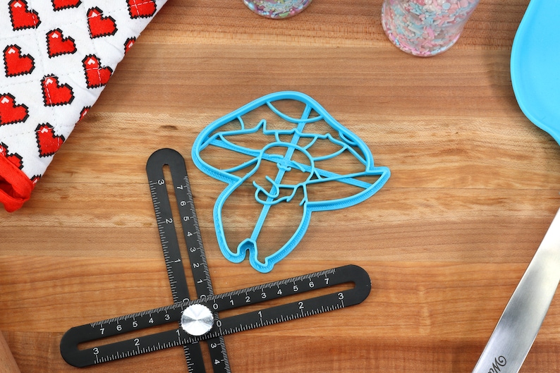 May include: A blue plastic cookie cutter shaped like a mushroom with a small mushroom on top. The cookie cutter is on a wooden surface with a ruler and other baking supplies.