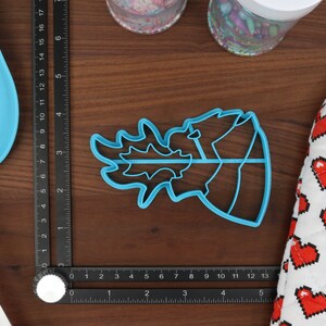 May include: A blue plastic cookie cutter shaped like a dragon's head with flames coming out of its mouth. The cutter is on a wooden surface with a ruler and a heart-patterned potholder.
