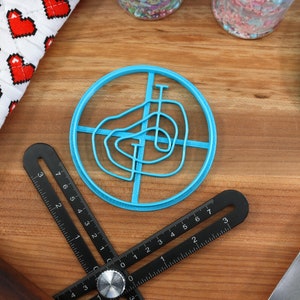 May include: A blue plastic cookie cutter shaped like a swirl design. The cutter is on a wooden surface with a ruler and a spatula.