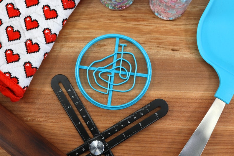 May include: A blue plastic cookie cutter shaped like a swirl design. The cutter is on a wooden surface with a ruler and a spatula.