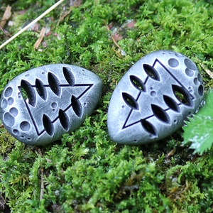 May include: Two gray stone pendants with a tree design carved into them. The pendants are on a bed of green moss.