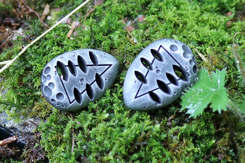 May include: Two gray stone pendants with a tree design carved into them. The pendants are on a bed of green moss.