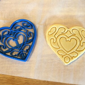 May include: A blue heart-shaped cookie cutter with a detailed design and a plain heart-shaped cookie on a white surface.