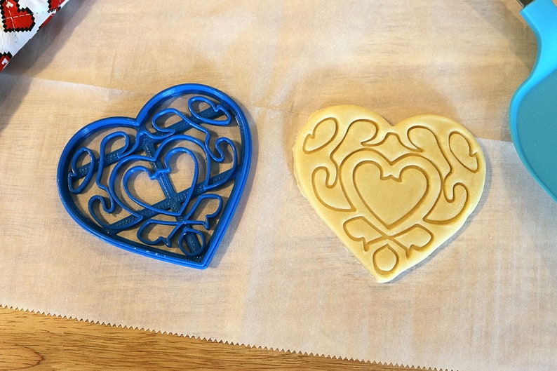 May include: A blue heart-shaped cookie cutter with a detailed design and a plain heart-shaped cookie on a white surface.