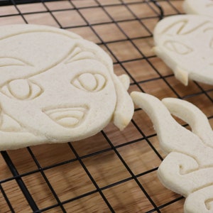 May include: Three white sugar cookies on a black wire rack. The cookies are decorated with simple designs, including a face with eyes and a mouth, and a swirl pattern.
