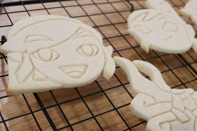 May include: Three white sugar cookies on a black wire rack. The cookies are decorated with simple designs, including a face with eyes and a mouth, and a swirl pattern.
