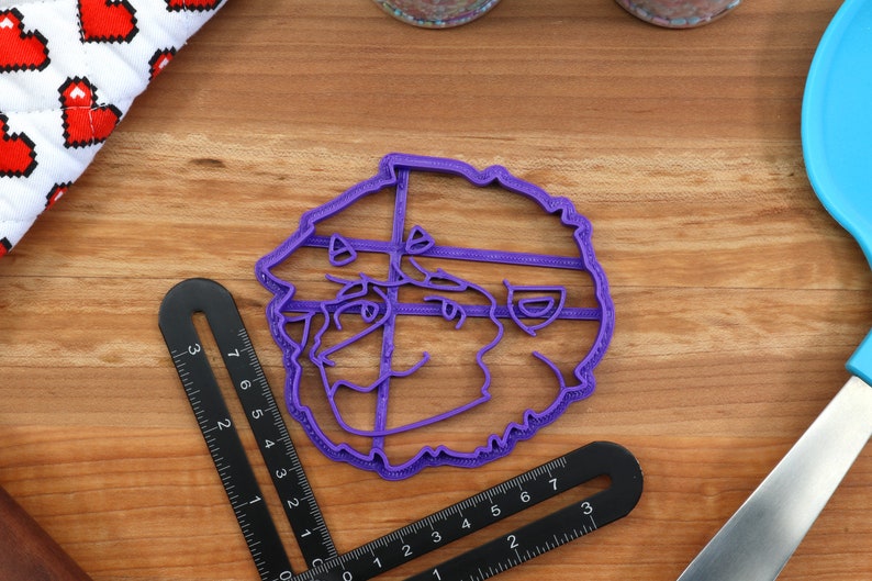 May include: Purple plastic cookie cutter shaped like a cartoon face with a wide smile and curly hair.