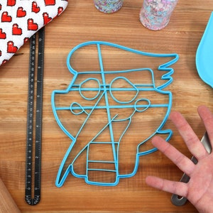May include: A blue plastic cookie cutter shaped like a cartoon character with a hat and a large smile. The cutter is on a wooden surface with a rolling pin, a ruler, and a spatula.
