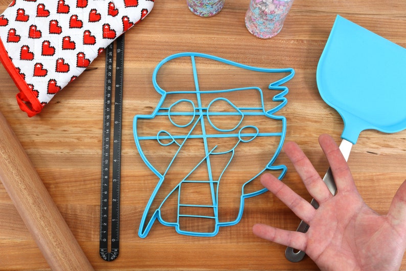 May include: A blue plastic cookie cutter shaped like a cartoon character with a hat and a large smile. The cutter is on a wooden surface with a rolling pin, a ruler, and a spatula.