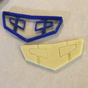 May include: A blue plastic cookie cutter in the shape of a stylized airplane wing sits next to a cut-out cookie of the same shape. The cookie and cutter are on a light-colored surface.