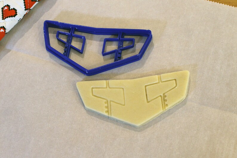 May include: A blue plastic cookie cutter in the shape of a stylized airplane wing sits next to a cut-out cookie of the same shape. The cookie and cutter are on a light-colored surface.