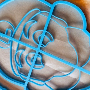 May include: A light blue plastic cookie cutter in the shape of a lion's head. The cutter has a detailed design with lines defining the facial features, including the mane, eyes, and nose. The cutter is placed on a light brown surface.