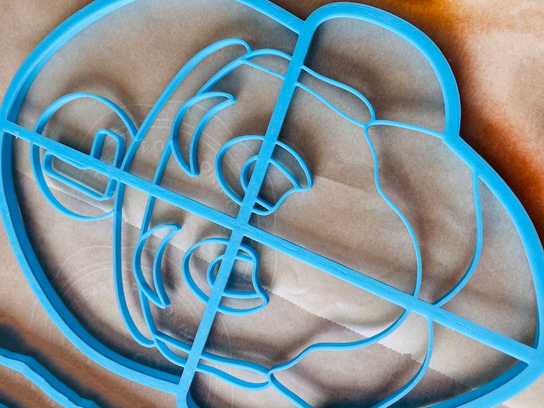 May include: A light blue plastic cookie cutter in the shape of a lion's head. The cutter has a detailed design with lines defining the facial features, including the mane, eyes, and nose. The cutter is placed on a light brown surface.