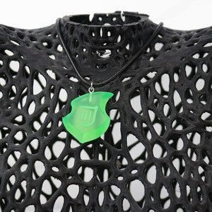 May include: A neon green pendant necklace with a shield design, suspended from a black cord. The pendant is displayed on a black, open-weave form. The shield features a stylized letter or symbol.
