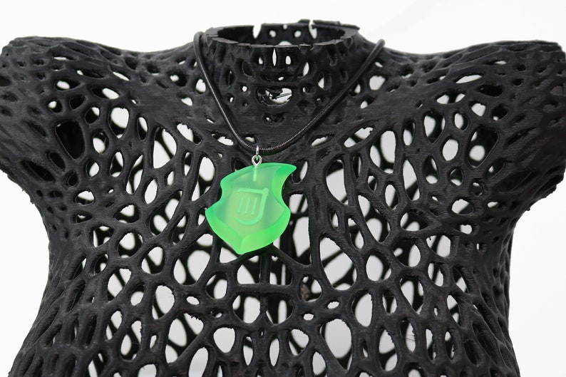 May include: A neon green pendant necklace with a shield design, suspended from a black cord. The pendant is displayed on a black, open-weave form. The shield features a stylized letter or symbol.