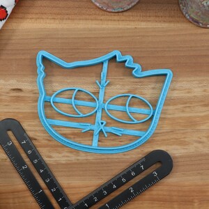 May include: Blue plastic cookie cutter shaped like a cat's face with large eyes and a nose.