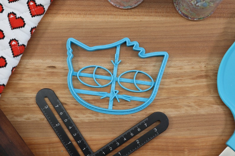 May include: Blue plastic cookie cutter shaped like a cat's face with large eyes and a nose.