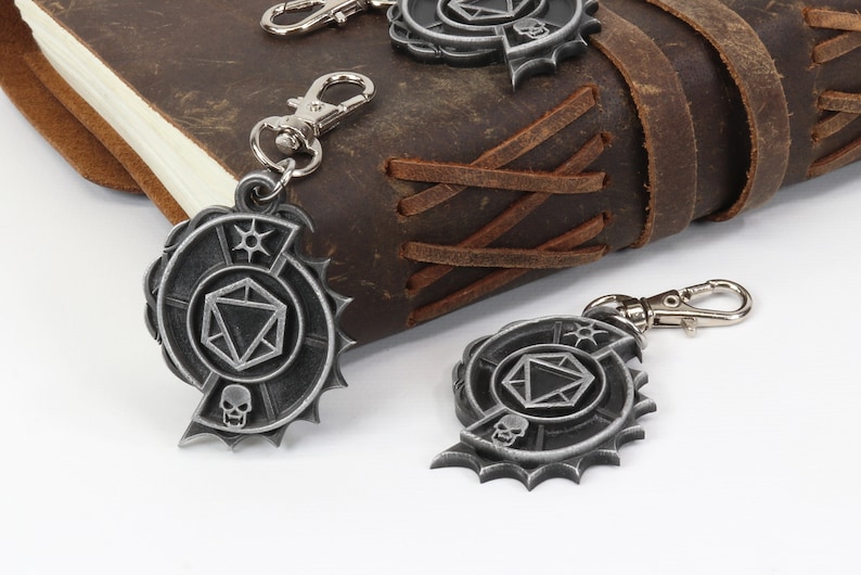 May include: Two silver metal keychains with a skull and a dice design. The keychains are hanging from a brown leather journal.