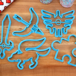 Puede incluir: Un juego de seis cortadores de galletas de plástico azul con forma de espada, arco y flecha, criatura alada, pieza de rompecabezas y escudo con un círculo en el centro.