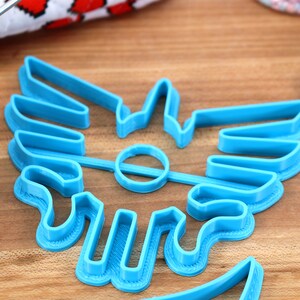 May include: A blue plastic cookie cutter shaped like a stylized winged symbol. The cutter is on a wooden surface.