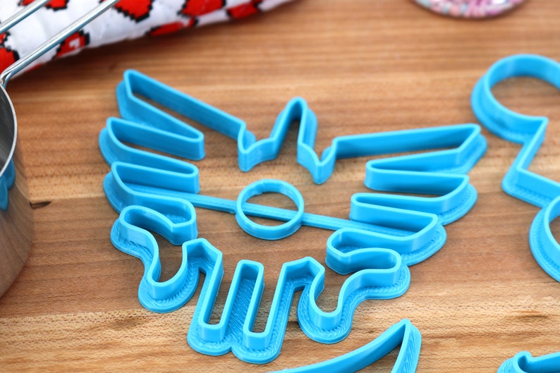 May include: A blue plastic cookie cutter shaped like a stylized winged symbol. The cutter is on a wooden surface.