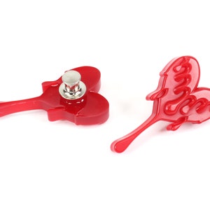 May include: Two red plastic heart-shaped buttons with a metal pin on one. The heart on the left has a raised design and the heart on the right has the word "Love" written in a script font.