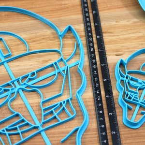 May include: Two blue plastic cookie cutters shaped like a cartoon character with a hat and glasses. The larger cutter is about 6 inches wide and the smaller cutter is about 3 inches wide.