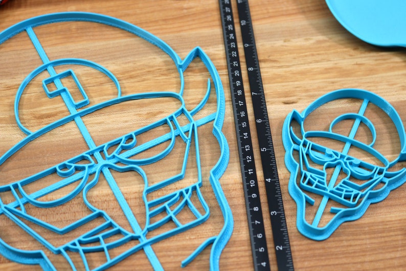 May include: Two blue plastic cookie cutters shaped like a cartoon character with a hat and glasses. The larger cutter is about 6 inches wide and the smaller cutter is about 3 inches wide.