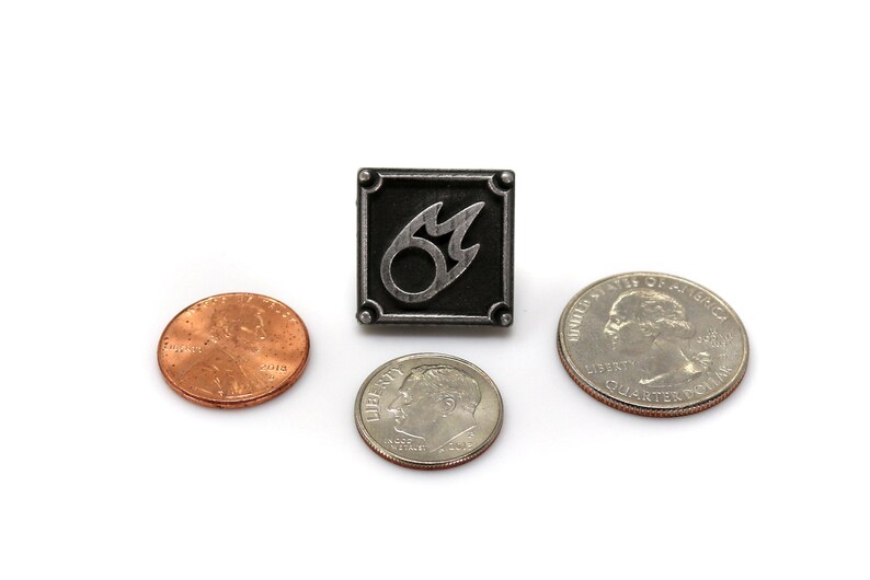 K&ouml;nnte beinhalten: Eine silberne Anstecknadel mit schwarzem quadratischem Hintergrund und einem silbernen Flammensymbol. Die Nadel ist von drei US-M&uuml;nzen umgeben: einem Penny, einem Dime und einem Quarter.