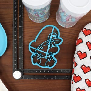 Op de afbeelding: Een blauwe plastic koekjesvormer in de vorm van een cartoonfiguur met een hoed. De koekjesvormer ligt op een zwarte liniaal met witte markeringen.