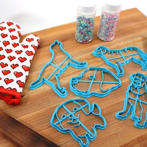 Peut inclure: Un ensemble de cinq emporte-pi&egrave;ces en forme de chien bleus sur une planche &agrave; d&eacute;couper en bois. Il y a aussi un gant de cuisine rouge, un rouleau &agrave; p&acirc;tisserie, une spatule bleue et deux pots de p&eacute;pites.
