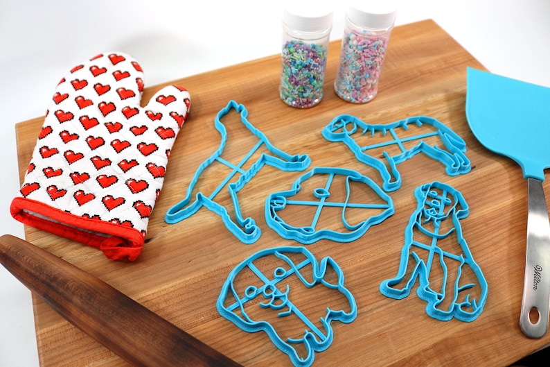 Peut inclure: Un ensemble de cinq emporte-pi&egrave;ces en forme de chien bleus sur une planche &agrave; d&eacute;couper en bois. Il y a aussi un gant de cuisine rouge, un rouleau &agrave; p&acirc;tisserie, une spatule bleue et deux pots de p&eacute;pites.