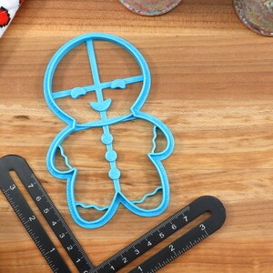 Op de afbeelding: Een blauwe plastic koekjesvormpje in de vorm van een gingerbread man. De vormpje heeft een glimlachend gezicht, knopen aan de voorkant en golvende lijnen aan de randen.