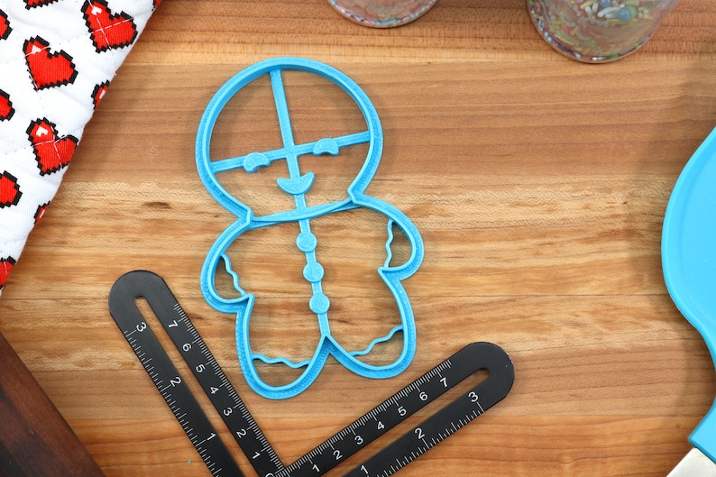 Op de afbeelding: Een blauwe plastic koekjesvormpje in de vorm van een gingerbread man. De vormpje heeft een glimlachend gezicht, knopen aan de voorkant en golvende lijnen aan de randen.